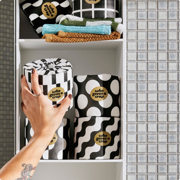 Who Gives A Crap premium toiler paper black and white packaging sitting on bathroom shelf with hand reaching to grab single roll