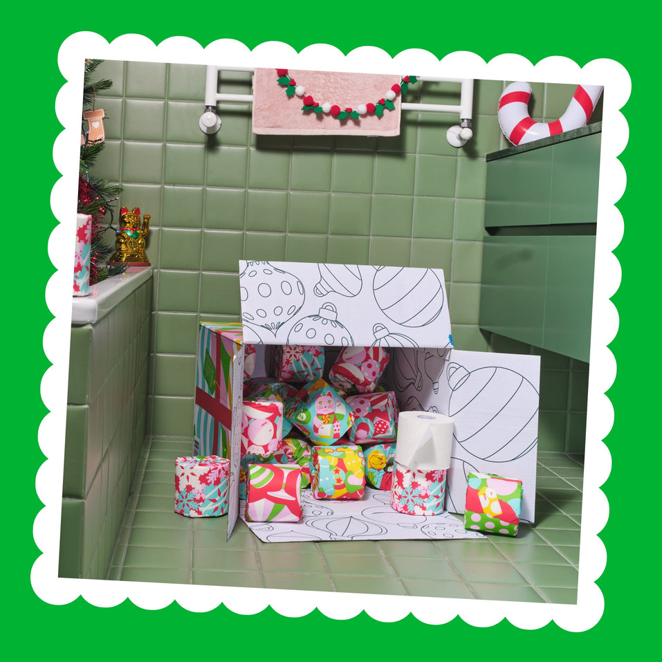 An open Gift Edition box overflowing with colourfully wrapped toilet paper rolls in a festive green-tiled bathroom, inside a scalloped white frame.