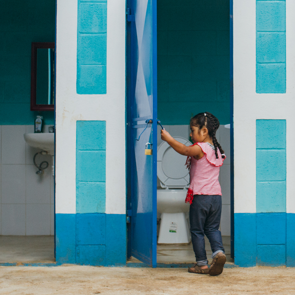 Young girl pushing open the door of a blue and white toilet block in Guatemala, a toilet and washbasin visible inside